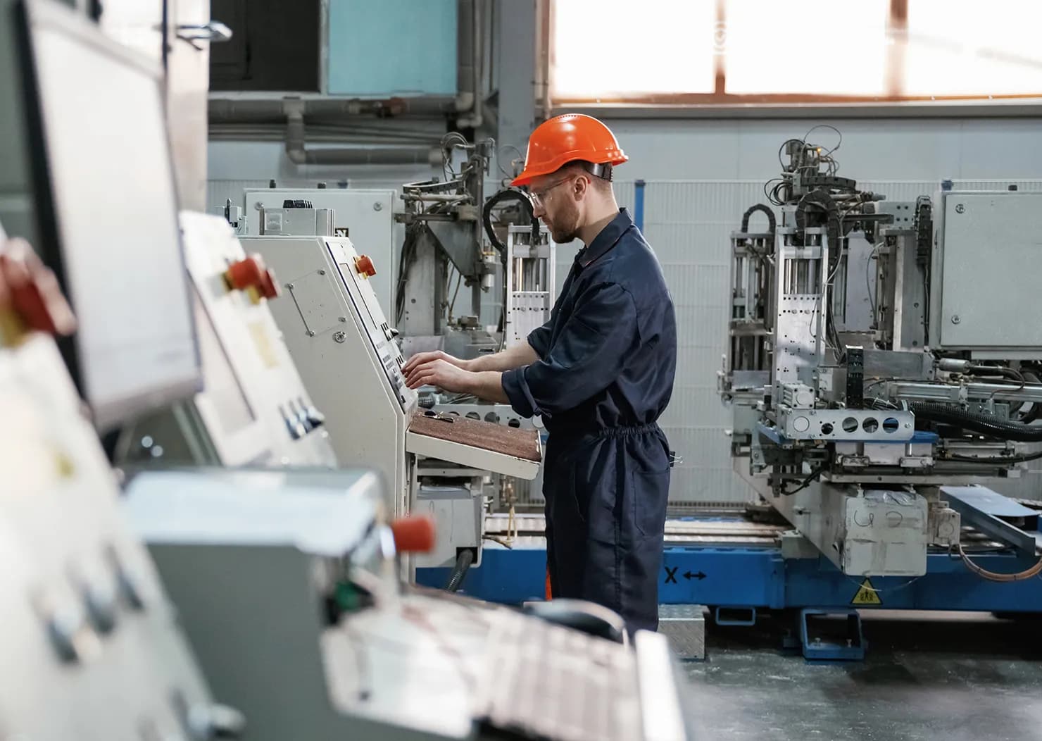 Pracownik ubrany w kask i kombinezon ochronny obsługuje panel sterowania w zakładzie produkcyjnym.