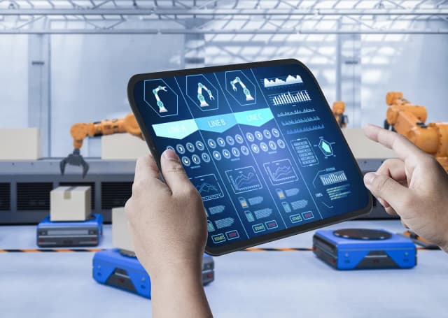 Osoba trzymająca tablet wyświetlający zautomatyzowane sterowanie fabryczne, ramiona robotyczne i roboty mobilne w magazynie